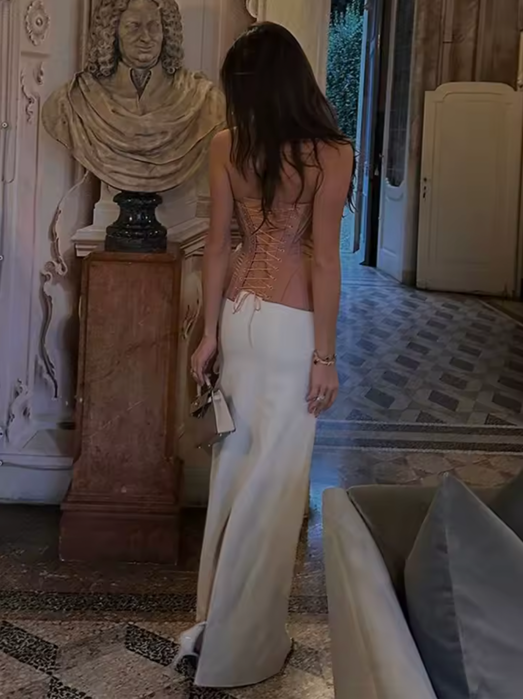 Woman in a beige top and white pants standing in a room with classical sculpture and patterned floor.