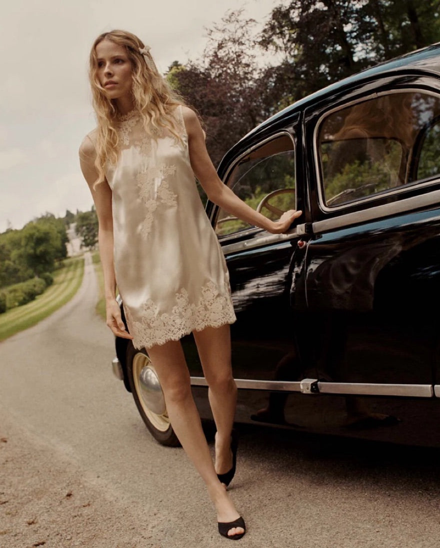 The Valeria Lace Dress in Candlelight combines buttery silk satin with refined 90s elegance. This sleeveless shift dress showcases a straight cut and a high neckline, offering a timeless silhouette. Its delicate lace details add a touch of playfulness, making it perfect for sophisticated occasions where subtle charm meets classic style.
