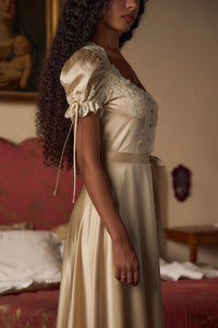 Crafted from luxurious silk charmeuse, the Unwrap Me Gown in Crème features delicate lace trim along the neckline and sleeves, enhancing its refined silhouette. Lace-covered buttons with hidden snaps offer elegant closure at the center front, while a matching silk charmeuse belt with chain stitch loops and snaps defines the waist. An adjustable slit secured by hook and thread loops adds both comfort and sophistication, making this gown a masterful blend of exquisite detail and expert tailoring.