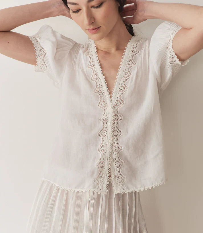 Crafted from lightweight semi-sheer ramie, the Brigitta Top by Doen draws inspiration from Victorian fashion with its plunging V-neckline and delicate lace detailing. The short puffed sleeves feature clustered pintucks, while a line of starburst buttons runs down the center front, adding refined charm. Versatile in style, it can be worn loose for an airy feel or tucked into high-waisted jeans for a polished look.