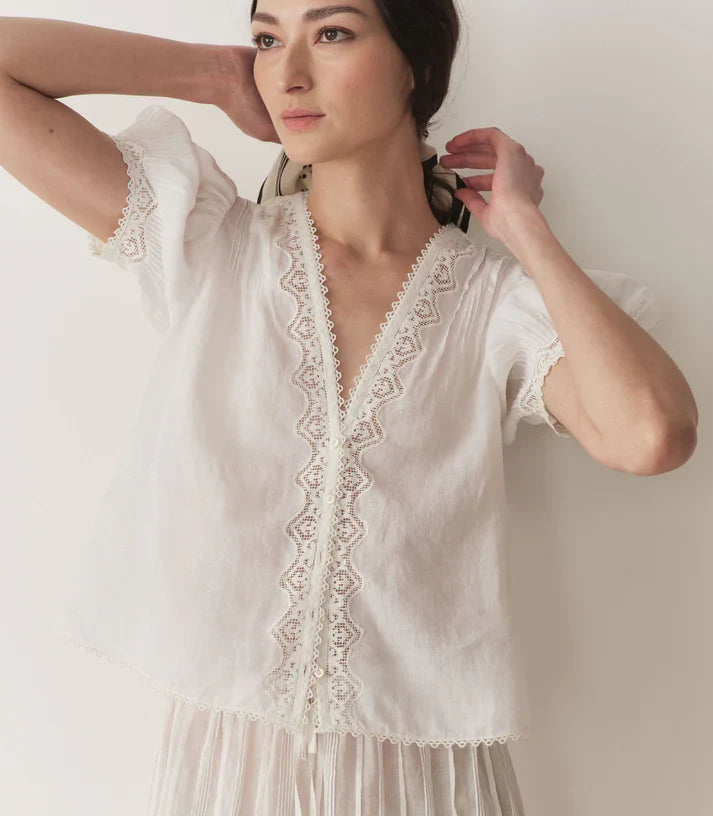 Crafted from lightweight semi-sheer ramie, the Brigitta Top by Doen draws inspiration from Victorian fashion with its plunging V-neckline and delicate lace detailing. The short puffed sleeves feature clustered pintucks, while a line of starburst buttons runs down the center front, adding refined charm. Versatile in style, it can be worn loose for an airy feel or tucked into high-waisted jeans for a polished look.