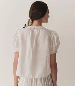 Crafted from lightweight semi-sheer ramie, the Brigitta Top by Doen draws inspiration from Victorian fashion with its plunging V-neckline and delicate lace detailing. The short puffed sleeves feature clustered pintucks, while a line of starburst buttons runs down the center front, adding refined charm. Versatile in style, it can be worn loose for an airy feel or tucked into high-waisted jeans for a polished look.