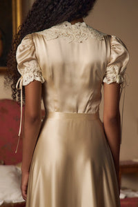 Crafted from luxurious silk charmeuse, the Unwrap Me Gown in Crème features delicate lace trim along the neckline and sleeves, enhancing its refined silhouette. Lace-covered buttons with hidden snaps offer elegant closure at the center front, while a matching silk charmeuse belt with chain stitch loops and snaps defines the waist. An adjustable slit secured by hook and thread loops adds both comfort and sophistication, making this gown a masterful blend of exquisite detail and expert tailoring.
