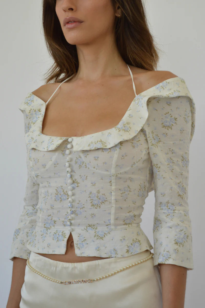Crafted from 100% organic cotton, the Margot Top in Vintage Iris by Fait Par Foutch offers a refined blend of comfort and elegance. Its adjustable lacing at the back ensures a tailored fit, complemented by delicate fold-over collar detailing and subtle non-functional buttons along the center front. Interior French seams enhance durability and a polished finish, while adjustable halter ties provide versatile styling options. 
