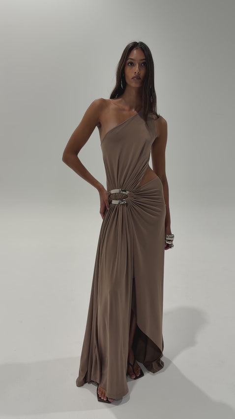The Serena Gown in Taupe is an elegant statement piece designed for those who appreciate sophisticated movement. A dramatic waterfall drape flows from one shoulder, creating a graceful frame that follows your silhouette. The design features a striking silver curved oval detail at the hip, adding a modern accent to the timeless taupe palette. A high split unveils an asymmetrical floating skirt that moves with natural ease, making every step feel effortless. 